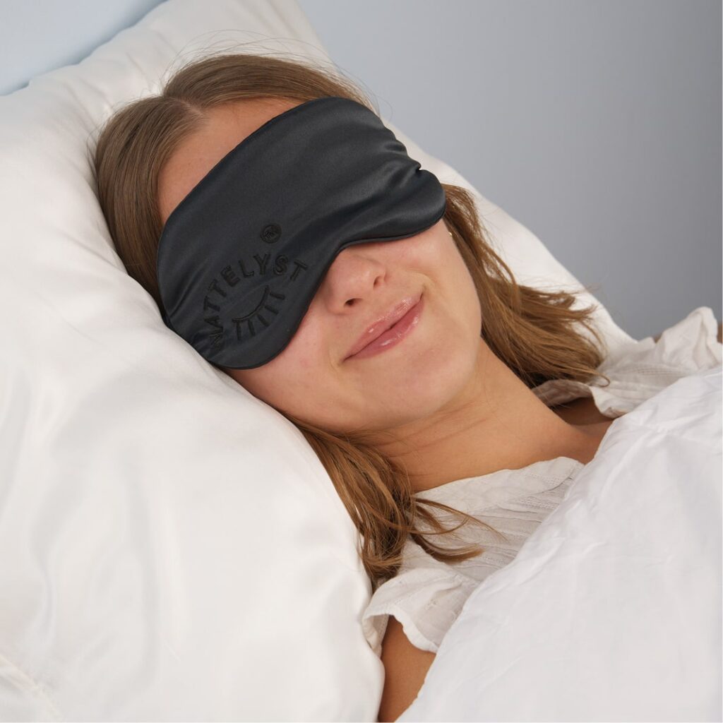 Smilende kvinde med sort sovemaske ligger afslappet i hvid seng