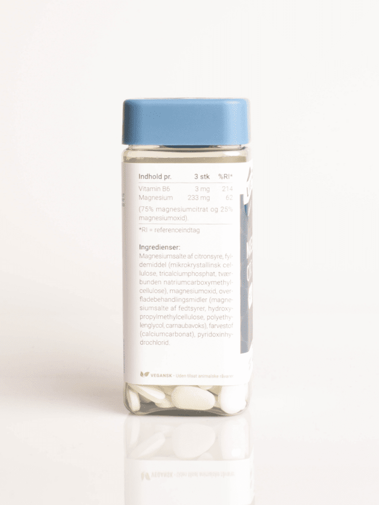 Klar glasflaske med kosttilskud, blåt låg og hvid etiket om magnesium og B6, synlige hvide tabletter