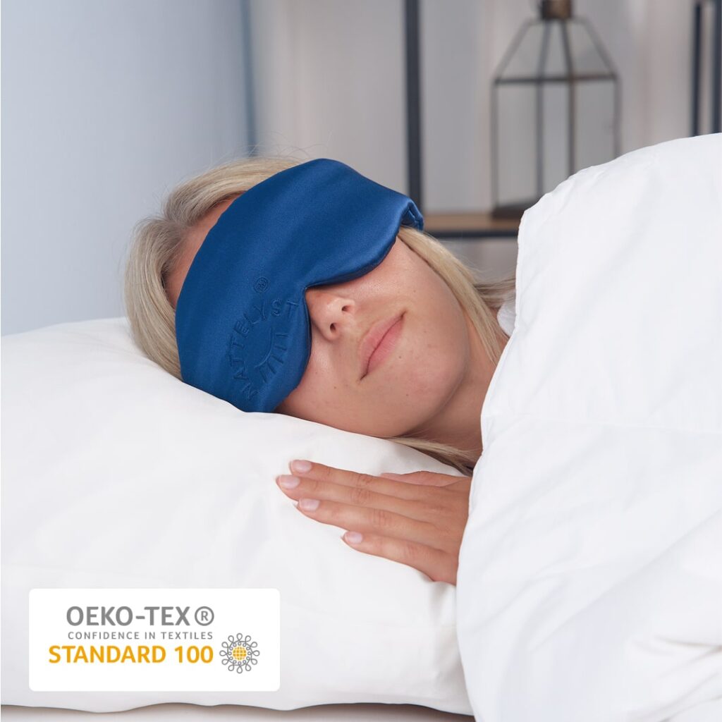 Kvinde sover med blød blå søvnmaske på hvidt sengetøj, OEKO-TEX Standard 100 mærke
